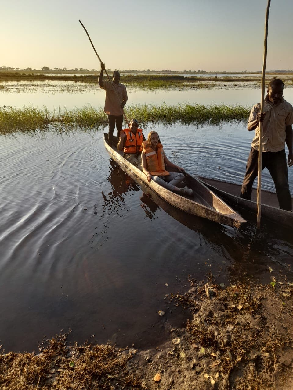 Two guides poling guests in a mokoro canoe through the shallows of the Chobe River at golden hour