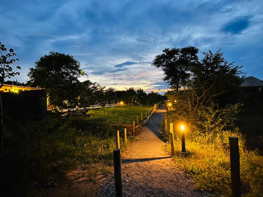 A lamp-lit gravel pathway through the lodge grounds at dusk, with the pool and loungers beyond